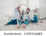 New building residential house purchase apartment concept. Stylish full family with two kids sitting on carpet, mom and dad making roof figure with hands arms over heads