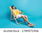 Full size photo positive guy enjoy rest relax exotic resort sun bathing hold glass cocktail sit deckchair wear white yellow striped shirt shorts barefoot isolated blue color background