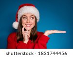 Photo positive girl in santa claus hat hold hand demonstrate x-mas newyear advert promo touch finger teeth wear warm winter season clothes isolated blue gradient color background