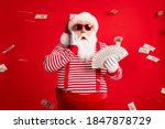 Photo of retired old man grey beard hold cash fan hand cheek shock money wear santa x-mas costume suspenders sunglass gloves striped shirt cap isolated red color background