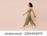 Side profile photo of cheerful nice woman with wavy hairdo dressed khaki overall white sneakers walking isolated on beige color background