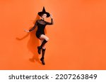 Full body photo of attractive young woman jump touch hat smile wear stylish black halloween witch look isolated on orange color background