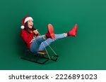 Full length photo of sweet funky girl dressed ugly print red x-mas pullover riding sledge looking empty space isolated green color background