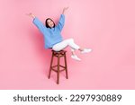 Full length photo of gorgeous pretty girl blue sweater white pants sitting on bar stool raising arms up isolated on pink color background