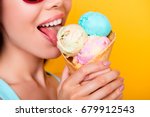 Close up cropped photo of young happy girl licking yummy ice cream of three scoops of different flavors, on yellow background, in sun glasses