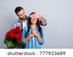 Handsome man with bristle in  shirt making  surprise for his beautiful, charming, joyful lover in casual outfit, closing her eyes with hands over grey background, women's day