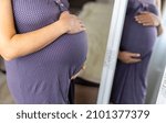 Small photo of Expecting female wearing a purple nightgown and her reflection. Caring future mom holds her belly tenderly. The tummy on the last months of pregnancy close up