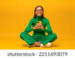 Small photo of Pretty millennial woman having fun laughing out loud sitting on floor with phone on yellow background browsing social media posts, laughing at funny memes, videos, stories ad reels