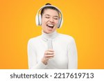 Teen student girl in white turtleneck wearing wireless headphones, singing loud in smartphone as microphone, having fun, isolated on yellow background