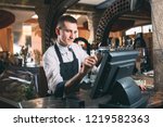 small business, people and service concept - happy man or waiter in apron at counter with cashbox working at bar or coffee shop