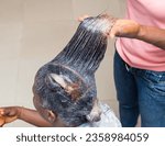 Small photo of Hand of an African stylist, holding, displaying and applying relaxer cream to the natural long hair of a woman or female customer at a beauty salon in Nigeria