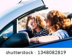 Two curly beautiful women friends drive and travel together on a convertible blue car having fun - outdoor happy leisure activity for cheerful people under the sun of summer - focus on second girl