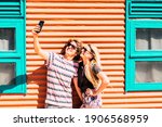 Small photo of Young modern cheerful couple man and woman take together a selfie picture in outdoor wirh colorful orange and blue wall in background - beautiful and youthful caucasian people enjoy sharing andhave fu