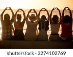 Group of women sitting against a golden sunset and doing heart symbol with hands and raised arms for friendship and love freedom concept. Women international day. Travel and vacation. Friends at beach