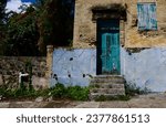 Small photo of an old dilapidated brick building with colorful door, steps, cracked concrete blue base, shuttered windows, weeds growing around the front stoop