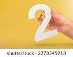 Small photo of Number two in hand. Hand holding white number 2 on yellow background with copy space. Concept with number two. 2 percent, birthday 2 years old, couple, two, double