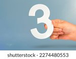 Small photo of Number three in hand. Hand holding white number 3 on blue background with copy space. Concept with number three. 3 percent, birthday 3 years, third grade, triple