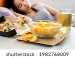 Small photo of Asian woman sleep after eating junk food with pizza, potato chips and glass of beer on desk, bad habit, unhealthy lifestyle concept
