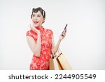 Small photo of Attractive gorgeous Asian woman wearing qi pao, cheongsam looking sideway holding shopping bags and phone exciting for sale event this lunar Chinese New Year season with copy space.