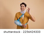 Handsome young asian man with broken arm in soft splint suffering a sore arm showing ok sign isolated on beige background, accident insurance concept.
