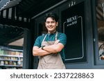 Small photo of Young adult Asian business man crossing her arms in front of cafe restaurant while smiling. Happy and joyful barista of Coffee shop having arms crossed. High quality photo