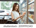 Small photo of Young beautiful Asian woman shopping and choosing items in supermarket. Female white collar worker picking something to drink at convenient store