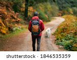 A hiker in a red coat and brown backpack walking with his white dog on a mountain path in spring or fall. Hiker, backpacker in autumn landscape. Mountain and trekking. Concept: Adventure, Art, Travel