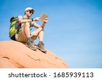 Hiker with backpack sitting outdoors using a tablet on the top of a red rock mountain