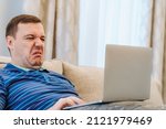 Small photo of Annoyed millennial man with disgust confused looking at laptop screen sitting on sofa at living room. Cringe guy looking at computer with squeamishness, reacting on watching disgusting video at home