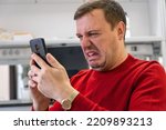 Small photo of Appalled businessman looking at phone screen sitting at workplace in lab office. Shocked man thinking of problem solution stuck with task. Confused guy reading news on phone with disgust