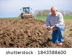 Smiling old-aged farmer kneeling and holding soil in his cupped hands with a tractor and a plough in the background