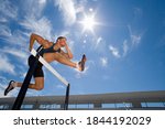 Small photo of Low angle view of a male athlete jumping over the hurdle with the background of sun flare and blue sky.