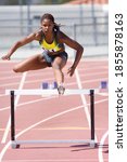 Small photo of Young African female runner jumping over a hurdle at an athletics event out at the track on a bright, sunny day