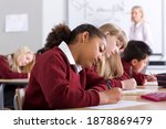 Sideview of�school children�sitting in�a�row�on desks�under�selective focus�as they take�a test�in�the�classroom�while the supervisor is�walking�in�the�background�