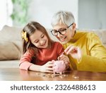 Small photo of Grandmother and her granddaughter Putting Coin Money In Piggybank At Home. Personal Savings, Bank Safety And Financial Investments Concept