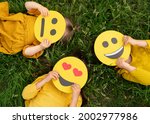 Three children lying on the grass are holding cardboard emoticons with different emotions in their hands: a sad, smiling happy smile, a loving smile with hearts instead of eyes.  World Emoji Day