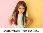 Small photo of Pretty cute charming girl with multicolored dyed strands of hair, isolated on a pink and yellow studio background. Safe coloring, hair care. Smooth and silky by nature. Female holds a lock of hair
