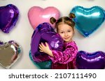 A cute three-year-old girl hugs a purple foil balloon in the shape of a heart. Decor photo zone of hearts for Valentine's Day