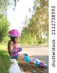 Small photo of Droll a five-year-old girl in a helmet, protective gloves, knee pads and roller skates sits on the curb of the sidewalk and laughs merrily. Summer sports entertainment