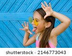 Small photo of A brunette girl in yellow sunglasses stands against a blue wall of planks and makes a funny grimace, showing her tongue. Naughty ill-mannered child