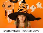 Small photo of An angry dissatisfied girl in the guise of a witch looks into the camera making a face. Halloween decor with ghosts on an orange studio background