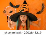 Small photo of A girl in the guise of a witch and a big black hat with a surprised face and an open mouth on an orange background with bats, cobwebs and spiders