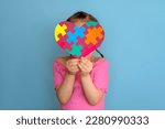 Small photo of A shy timid girl in a pink T-shirt covers her face with a heart made of colorful puzzles. To tell , inform and educate to be tolerant to people suffering from autism spectrum disorder