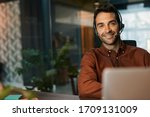 Small photo of Portrait of a smiling businessman talking to a client over a headset, while working on a laptop at his desk in a dark office after hours