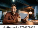 Small photo of Portrait of a smiling businessman sitting after hours alone at his dark office desk and working online with a digital tablet