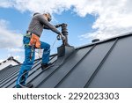 Small photo of Work at heights. A tinman with tool belt and climbing harness on the roof is working with a cordless drill on the chimney sheeting.
