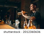 Young handsome smiling male bartender in apron preparing alcohol drink shaking it in cocktail shaker standing in front of bar counter. Side view