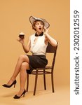 Small photo of Gossip, tattle. Pretty woman wearing elegant clothes with hat holding glass of beer and talking via cell phone over beige background. Concept of drinks, alcohol, vacation, party, celebrating, ad