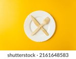 Small photo of Wooden fork and knife lie on a white plate crosswise. Tableware from natural materials isolated on a yellow background. Top view. Plastic free concept. Diet concept.
