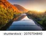 Aerial view of silhouette of walking man on the suspension bridge, river, mountains, red trees, green grass, orange sun at sunset in autumn. Colorful landscape with forest, lake, reflection in water